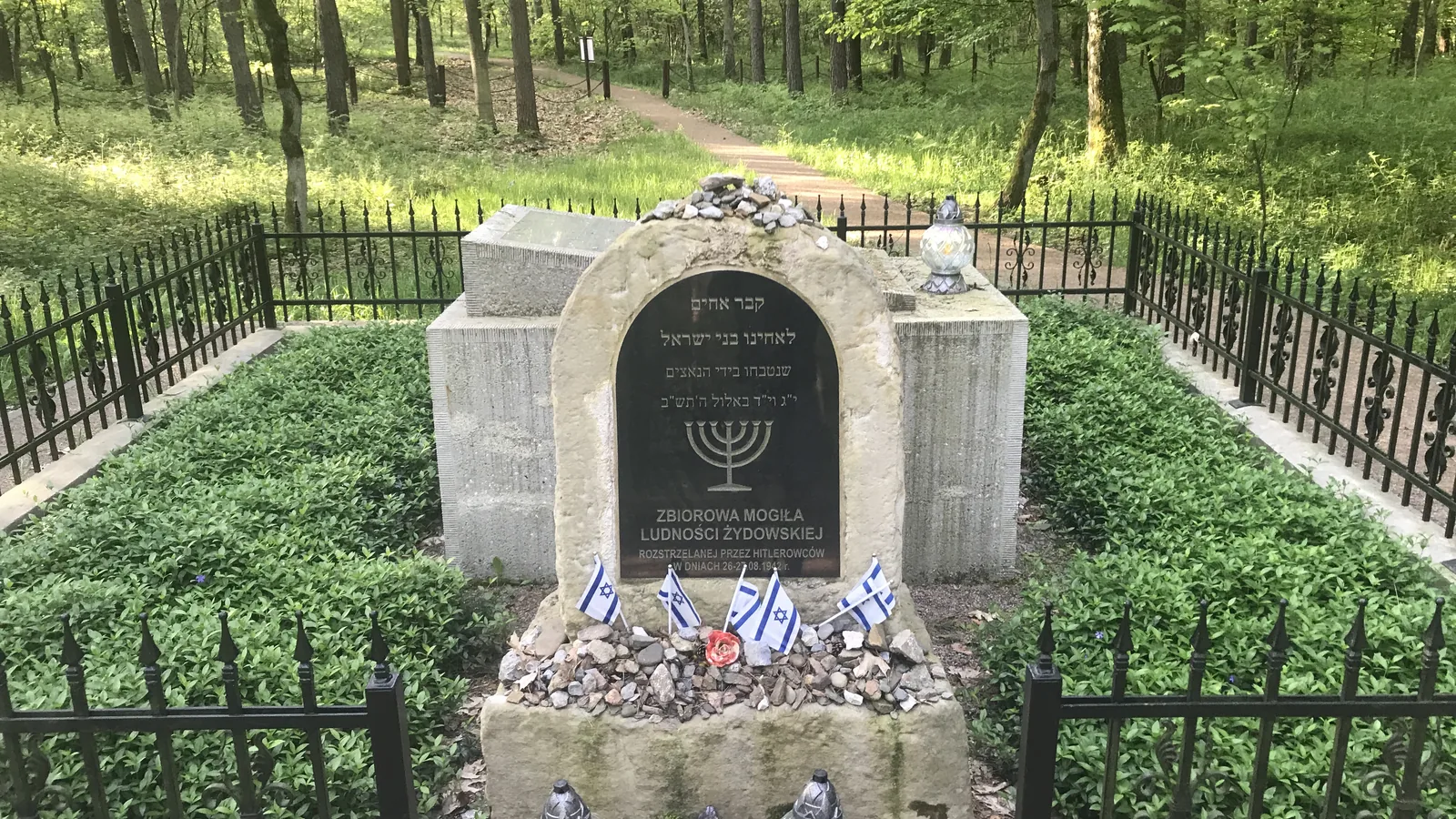 Collective grave of the Jewish community in a forest - a matzeva with a menorah, pebbles and small Israeli flags, surrounded by a fence