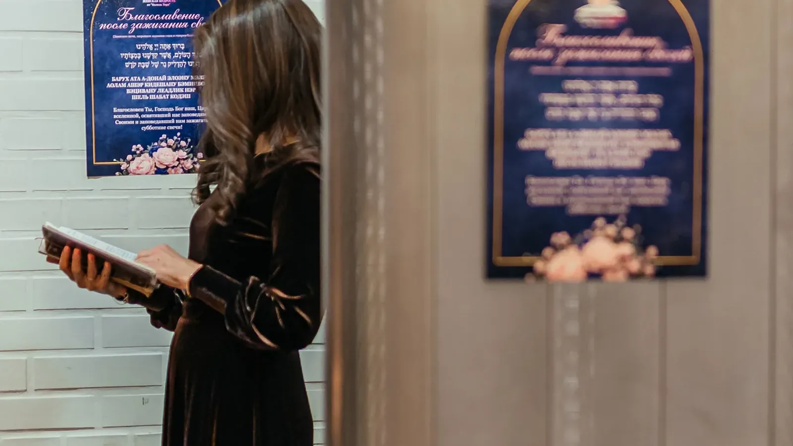 A woman reading a prayer from a siddur beside a plaque with the blessing after candle-lighting